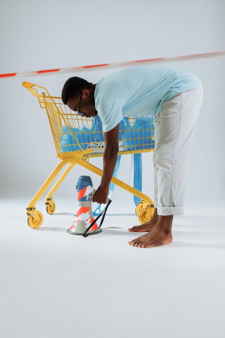 Barefoot man with a megaphone near a yellow shopping cart in a minimalistic indoor setup.